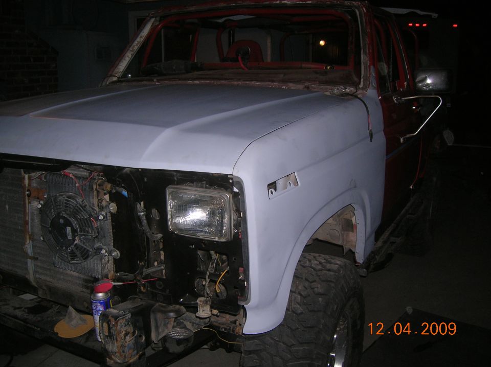Front end of a Ford Bronco with primer on the hood and fender, missing grille and headlights. Roll cage visible inside