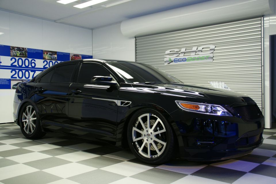 Black sedan with custom wheels parked indoors on a checkered floor. Wall displays "2006 2007" timeline