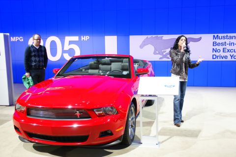 1993 Ford Taurus - A red convertible Mustang is displayed at an auto show with a speaker presenting.