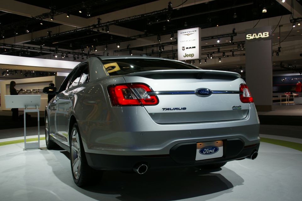 Rear view of a silver Ford Taurus at an auto show. Shows 'TAURUS' badge and 'Ford' license plate frame