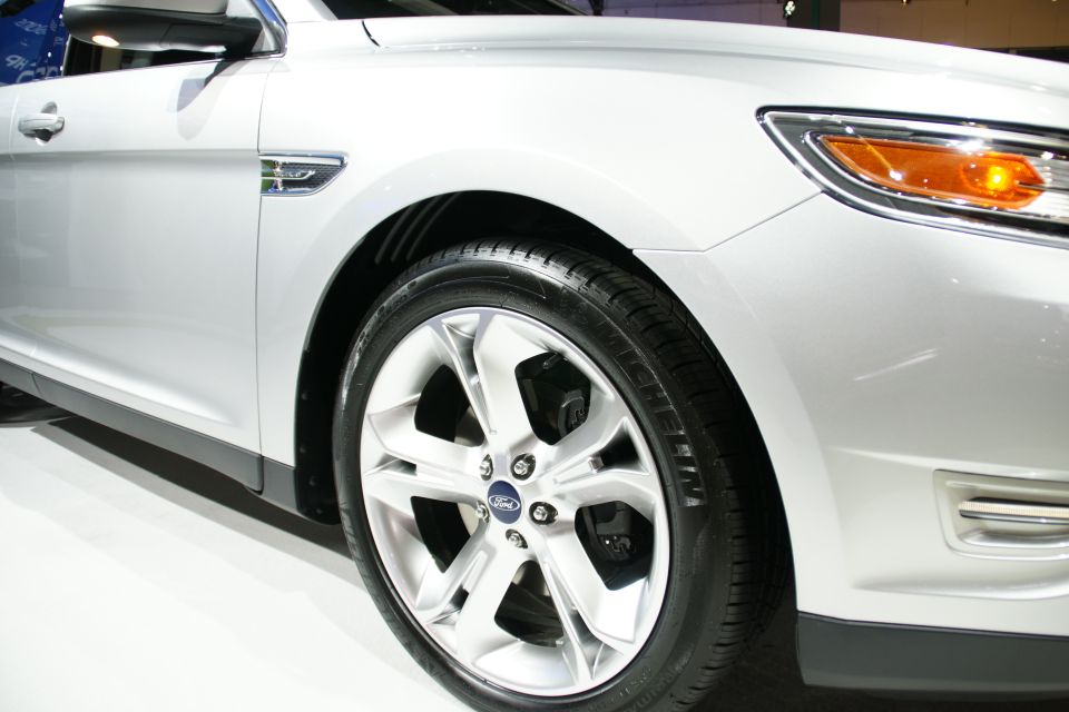 Close-up of a silver sedan's front wheel and fender. Shows a Ford logo on the wheel