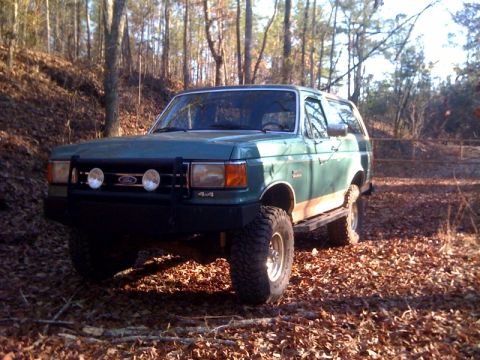 1989 Ford Bronco - Green 1989 Ford Bronco with aftermarket bumper and lights in a wooded area.