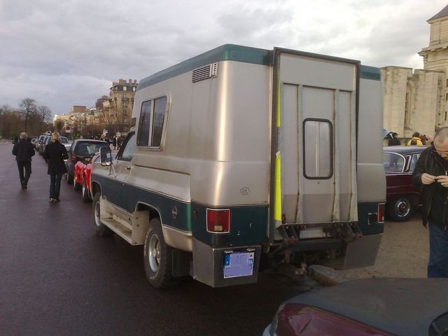 Camper truck parked in a line of cars, with a building in the background