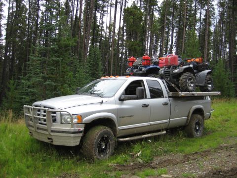 2005 Dodge 3500 - A silver Dodge Ram 3500 pickup truck is loaded with two ATVs and fuel cans, parked on a muddy track in a wooded area.