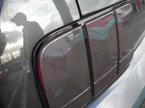 1998 Ford Crown Victoria - Close-up of a gray vehicle's rear quarter panel and taillight, reflecting a person wearing a baseball cap.
