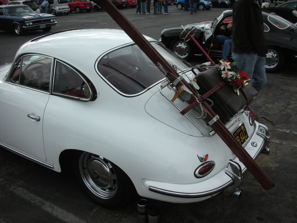 White Porsche 356 with skis and suitcase strapped to rear rack. Other cars and people in background