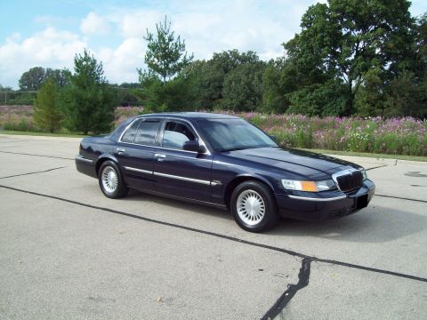 1999 Mercury Grand Marquis - Dark navy blue Mercury Grand Marquis full-size sedan photographed from the front three-quarter angle on an empty concrete lot. The car wear…