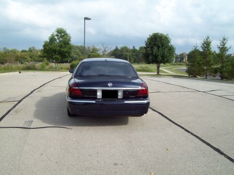 1999 Mercury Grand Marquis - Straight-on rear view of a dark navy blue Mercury Grand Marquis sedan in an empty crack-sealed parking lot, showing the full-width chrome t…