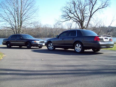 1999 Mercury Grand Marquis - Two dark sedans are parked on an asphalt surface near a body of water, with trees in the background.