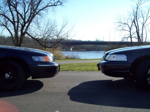 1999 Mercury Grand Marquis - Two sedans are parked facing each other near a body of water with a bridge in the background.