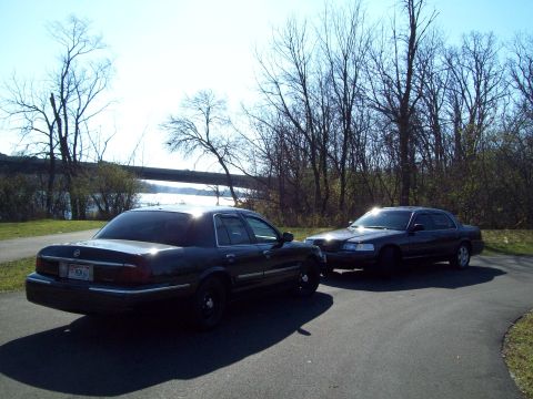 1999 Mercury Grand Marquis - Two dark sedans are parked on a paved path with trees and a body of water in the background.
