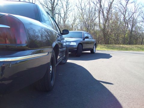 1999 Mercury Grand Marquis - Two dark sedans, one a 1999 Mercury Grand Marquis, are parked side-by-side on an asphalt surface with trees in the background.