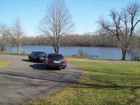 1999 Mercury Grand Marquis - Two sedans are parked on a paved area next to a body of water on a clear day.