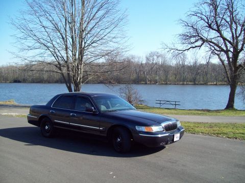 1999 Mercury Grand Marquis - A dark sedan is parked on the side of a road next to a lake on a clear day.
