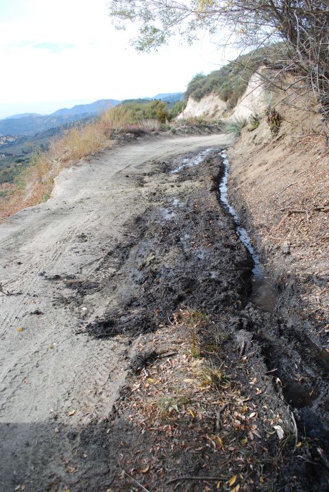 1996 Ford Bronco - A muddy dirt road winds up a hillside, with water runoff carving a path alongside it. Distant mountains are visible under a hazy sky.