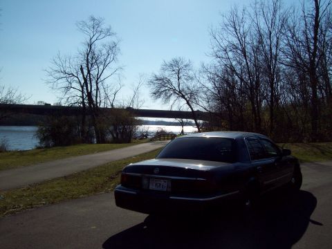 1999 Mercury Grand Marquis - A dark sedan is parked on a path next to a body of water with trees and a bridge visible.