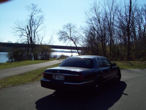 1999 Mercury Grand Marquis - A dark sedan is parked near a body of water with trees in the background.