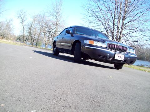 1999 Mercury Grand Marquis - A dark sedan is parked on the side of a road near a body of water. The license plate is visible.