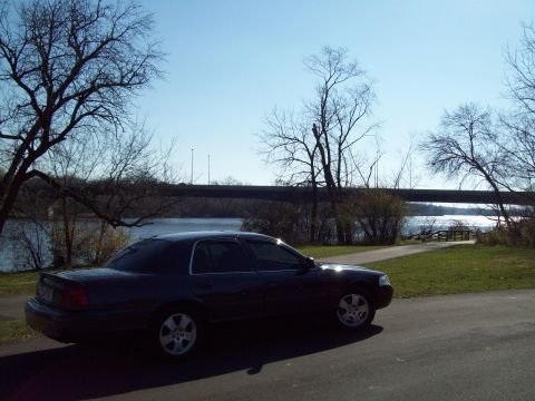 1999 Mercury Grand Marquis - A dark sedan is parked on a paved surface next to a grassy area with trees and a river.