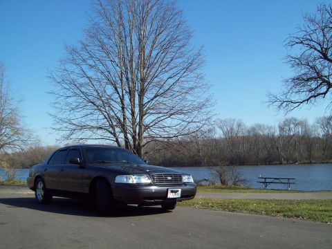 1999 Mercury Grand Marquis - A dark sedan is parked by a lake on a clear day, with bare trees and a picnic table visible.
