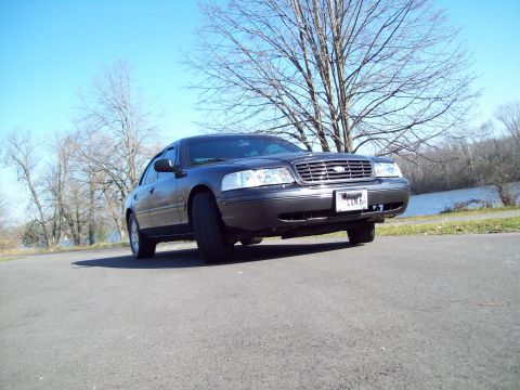 1999 Mercury Grand Marquis - A dark sedan is parked on an asphalt surface with a body of water and trees in the background. The license plate is visible.