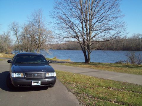 1999 Mercury Grand Marquis - Dark sedan parked on a paved path next to a body of water on a clear day.