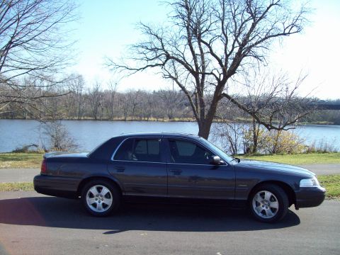 1999 Mercury Grand Marquis - A dark sedan is parked on a paved surface next to a calm body of water under a bright sky.