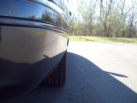 1999 Mercury Grand Marquis - A dark sedan's rear quarter panel and tire are visible from the side, with trees in the background.