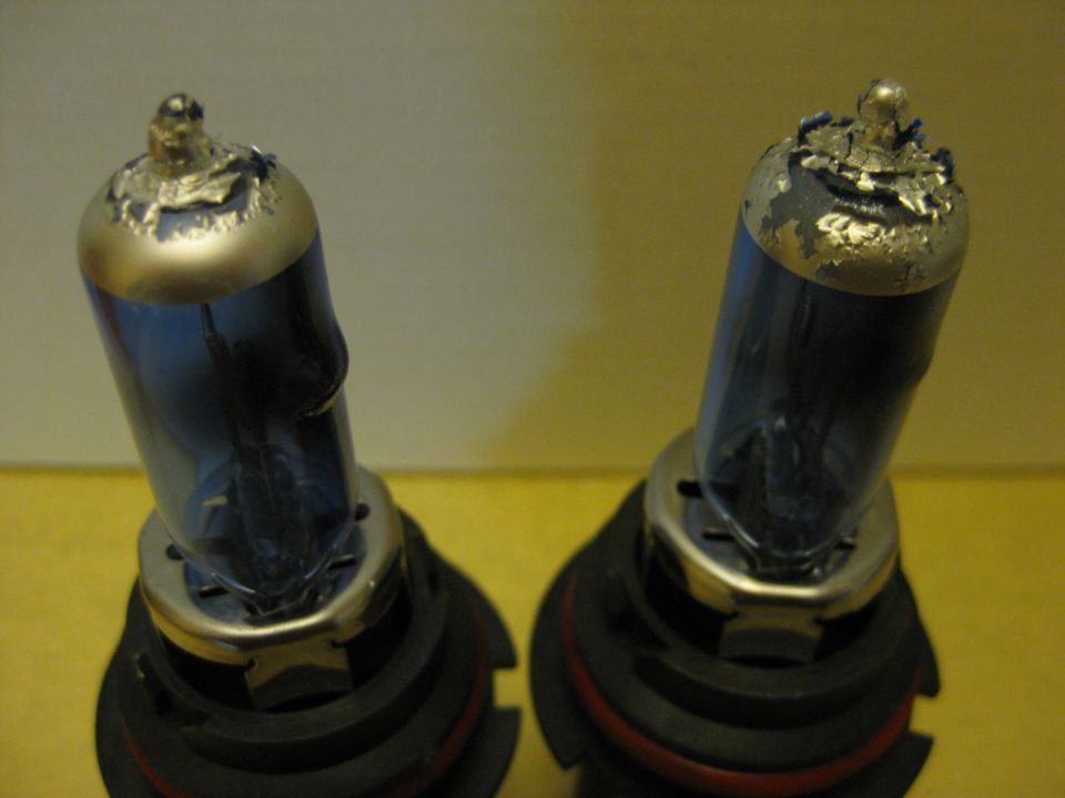 Two blue-tinted headlight bulbs with damaged tops sit on a yellow surface. Supermotors.net logo visible