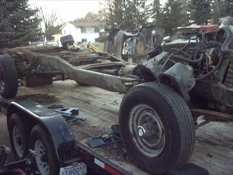 Truck chassis and engine assembly on a flatbed trailer. The frame is heavily rusted