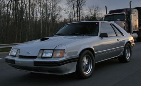 1986 Ford Mustang - A silver 1986 Ford Mustang coupe is seen on the highway, with a semi-truck visible in the background.