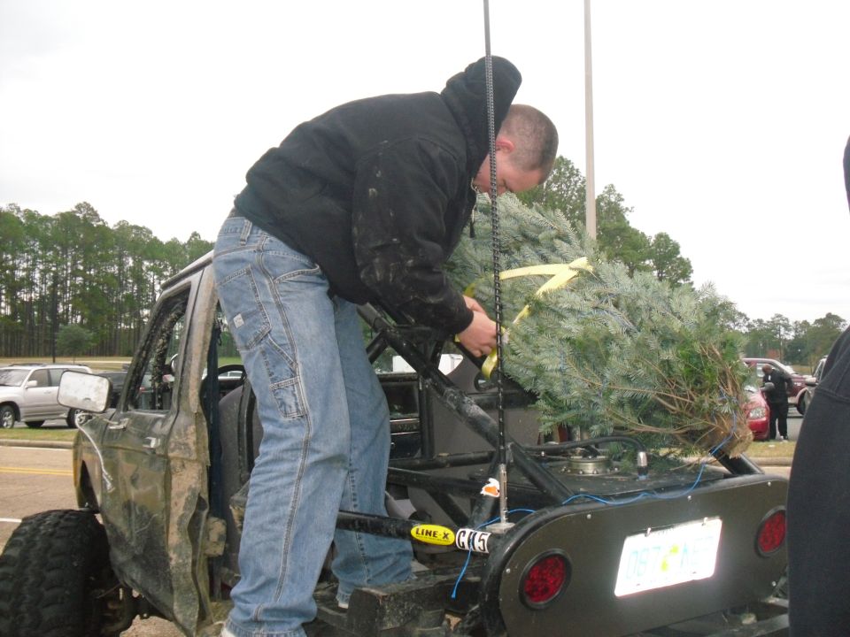 Guy securing a Christmas tree to the back of a modified off-road SUV. Another SUV is visible in the background
