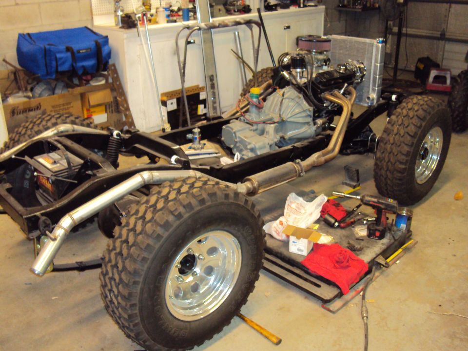 Jeep CJ chassis with engine, transmission, and radiator installed. Tools and parts are on a cart nearby