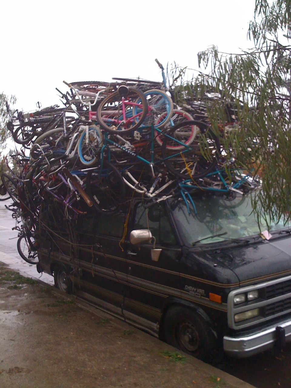 Black Chevrolet van with a massive pile of bicycles strapped to its roof, parked on a dirt shoulder