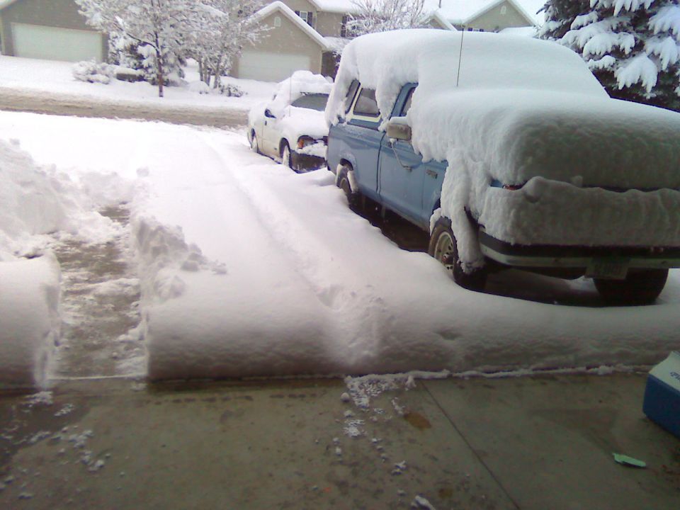 Blue pickup truck and white car parked in a driveway, both covered in a thick layer of snow. A path has been shoveled