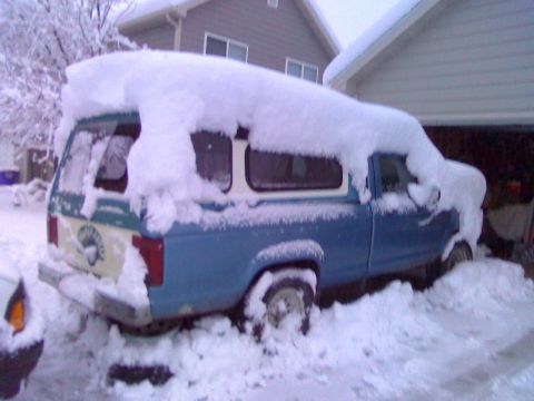 2006 Ford E350 - A blue pickup truck is partially buried in snow, with a camper shell on its bed, parked near a garage.