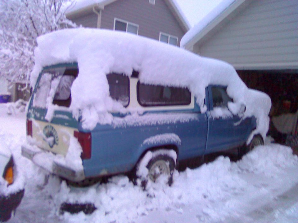 Blue pickup truck covered in snow, parked next to a garage. A camper shell is on the bed