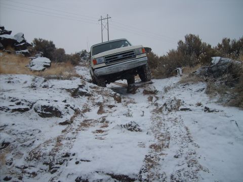 1989 Ford Bronco - A white Ford truck drives through a snowy, off-road terrain with rocky obstacles.