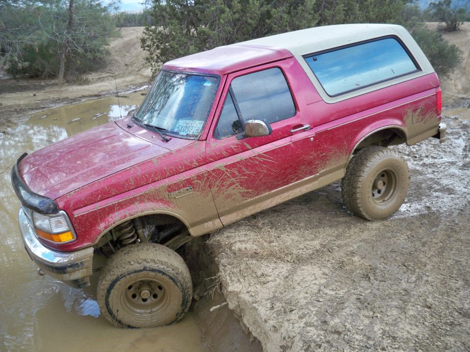 Red Ford Bronco with a tan top stuck in mud, front left wheel submerged