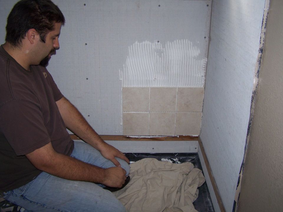 Man in brown shirt and jeans tiling a wall with tan tiles