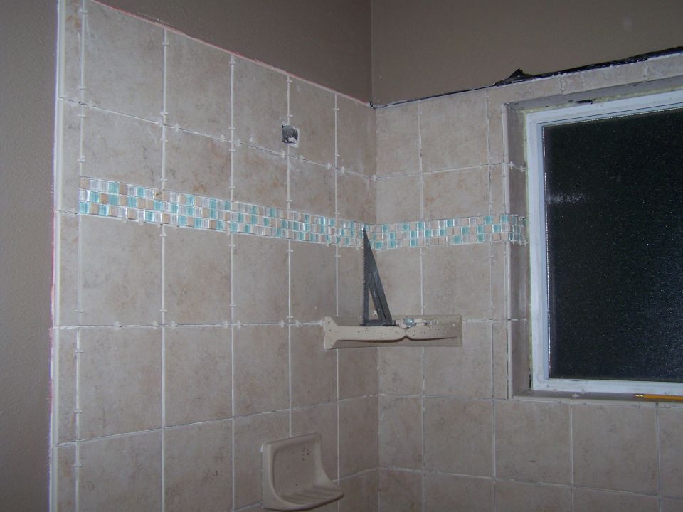 Bathroom corner with tan tiles and a mosaic accent strip; window on right. Tiling is incomplete