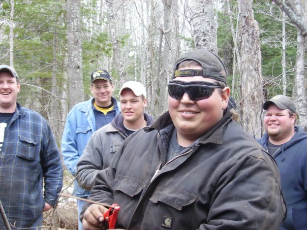 Five men stand in a wooded area. The man in the foreground wears sunglasses and a baseball cap
