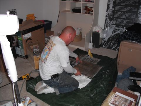 1991 Ford Bronco - A man kneels on a tarp, using a tile cutter to prepare flooring materials amidst a room filled with boxes and supplies.