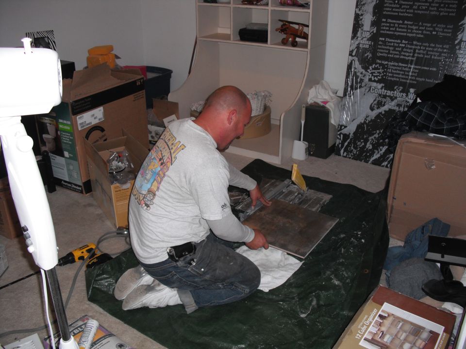 Man cutting tile on a tarp indoors, surrounded by boxes and home improvement supplies