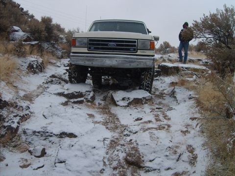 1989 Ford Bronco - A white Ford F-Series truck navigates a challenging, rocky path covered in snow.