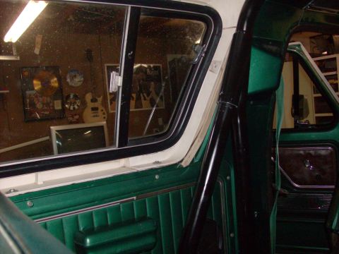 1978 Ford Bronco - Interior view of a green vehicle, with a roll cage and reflections of music memorabilia in the window.