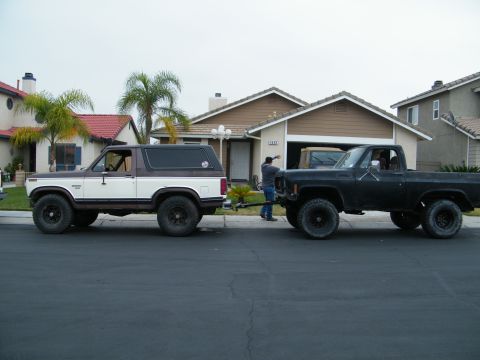 1982 Ford Bronco - A two-tone Ford Bronco from the early 1980s is towed by a black pickup truck on a suburban street.