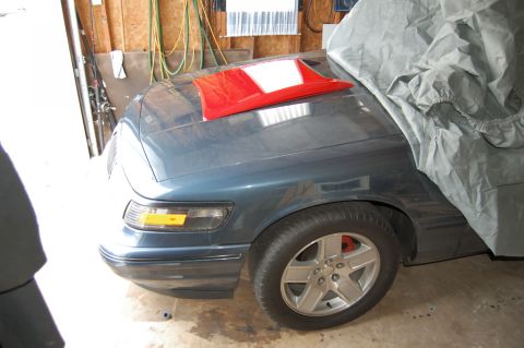 1985 Mercury Grand Marquis - A red hood scoop is placed on the hood of a blue car, which is partially covered by a car cover in a garage.