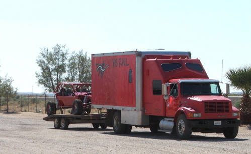 Red box truck towing a flatbed trailer with a red sand rail buggy on gravel.