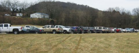 2007 Ford Mustang - A variety of trucks and cars are parked in a line in a grassy field with a wooded hill in the background.